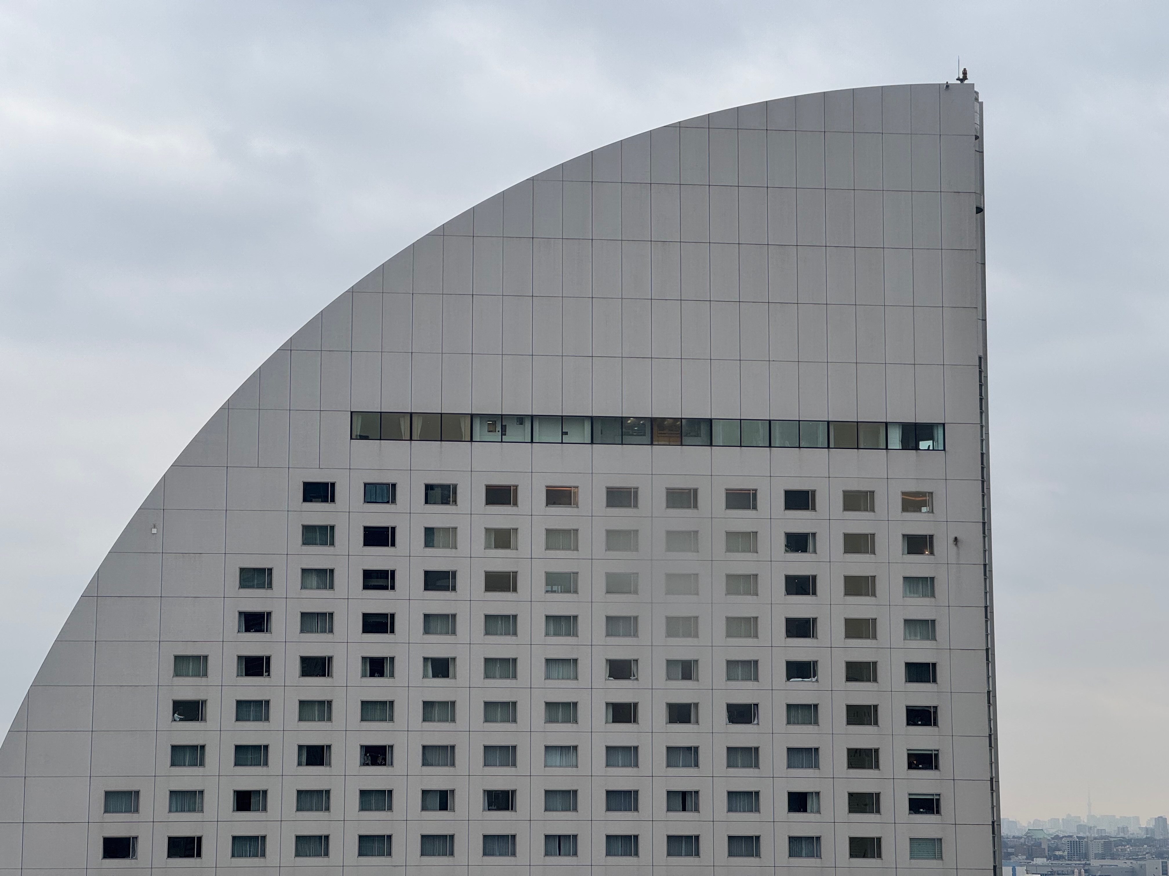 A large white building with a curved top and numerous windows stands tall against a cloudy sky, with a person perched on a balcony on the top floor.