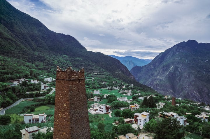 A serene mountain town with white buildings and green roofs is nestled amidst lush greenery, surrounded by towering red and green mountains. A tall brick tower stands in the foreground, contrasting with the natural surroundings. The sky is filled with clouds, casting a soft light over the scene.