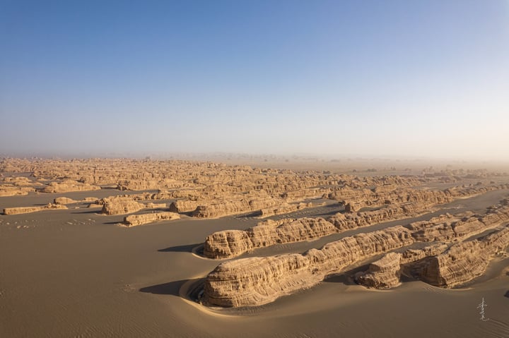 Aerial view of a desert landscape with sandy formations, a clear blue sky, and a few distant structures.