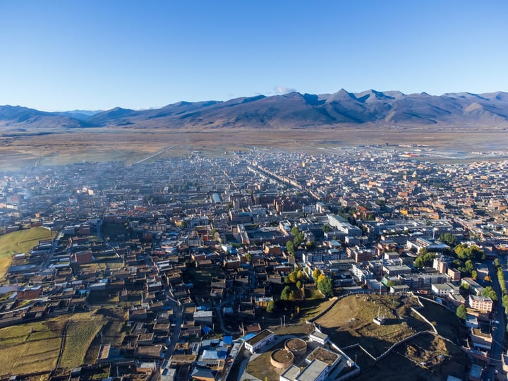 An aerial view of a city nestled between mountains, with a mix of buildings and green spaces, surrounded by snow-capped mountains.