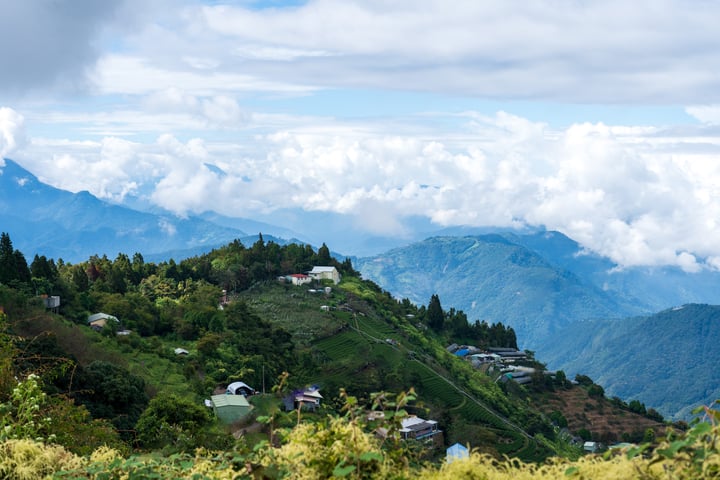 A serene mountainous landscape with houses nestled among lush greenery, surrounded by towering mountains and a clear blue sky with fluffy white clouds.