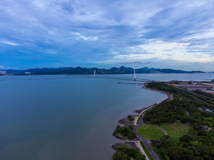 An aerial view of a large body of water with a bridge spanning across it, surrounded by lush greenery and mountains. The sky is filled with clouds, and the water is calm.
