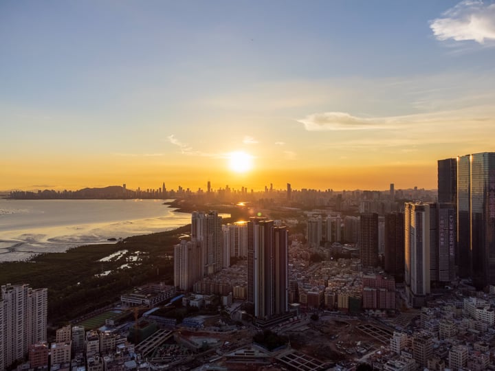 An aerial view of a bustling city at sunset, with tall buildings reflecting the setting sun, a river flowing through the city, and a large body of water in the foreground.