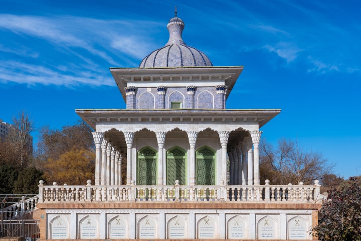 A white, ornate gazebo with a dome and a small tower is situated on a hill, surrounded by lush greenery and a clear blue sky.