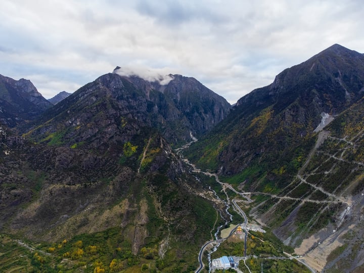 An aerial view of a mountainous valley with a winding river, surrounded by towering mountains and greenery, with a cloudy sky overhead.