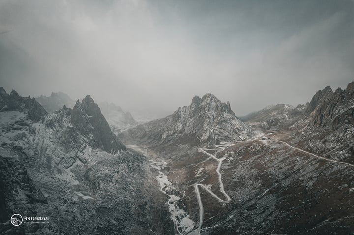 A snowy mountain range with a winding road through it, surrounded by foggy mountains and a cloudy sky.