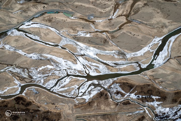 An aerial view of a river winding through a sandy landscape, with a light blue river and a darker blue river in the background, creating a sense of depth and vastness. The image is taken from above, providing a bird's eye view of the landscape.