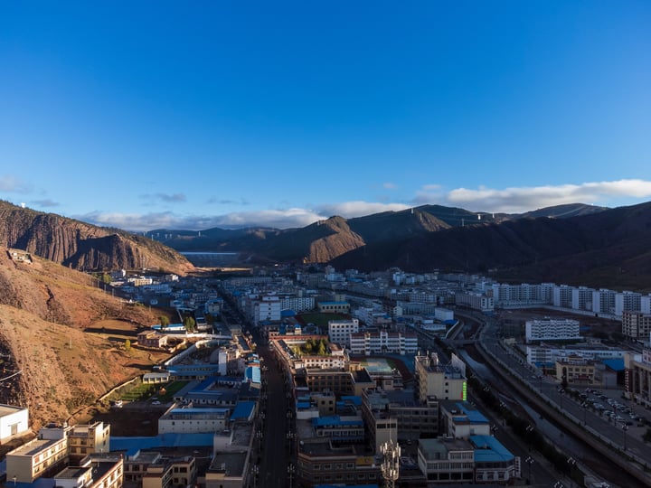 A cityscape with a mix of buildings and roads, surrounded by mountains and a clear blue sky.