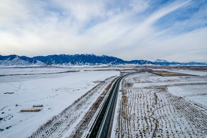 An aerial view of a snow-covered road leading to mountains, with a clear blue sky above.