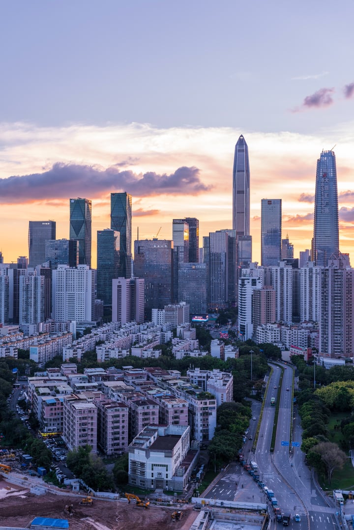 A cityscape at sunset, with tall buildings in various colors and shapes, a road with construction equipment, and a sky of orange and pink hues.