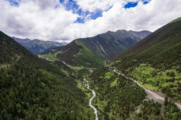 An aerial view of a winding mountain valley with a winding river, surrounded by lush green mountains and a clear blue sky with white clouds.
