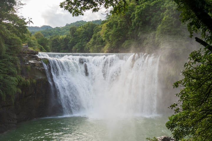 A waterfall cascades down a cliff, surrounded by lush greenery and a clear blue body of water.