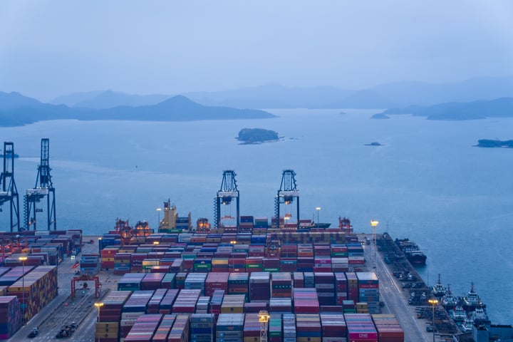 A bustling port with colorful shipping containers, cranes, and boats, surrounded by mountains and a large body of water.