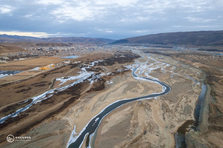 An aerial view of a winding river in a valley, surrounded by sandy and brown hills, with buildings and mountains in the distance.