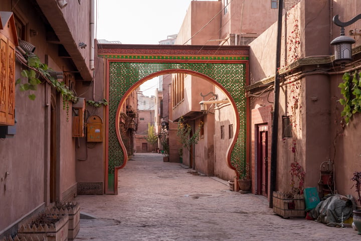 A narrow cobblestone street in a traditional Kashgar is lined with pink and brown buildings, some with green and red accents. An archway with green and red patterns leads to a red door, and potted plants are scattered around. The sky is clear, and the sun shines brightly.