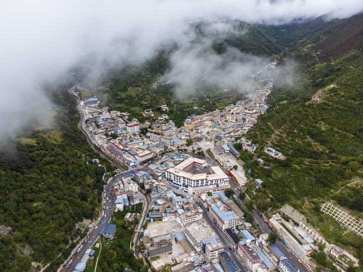 An aerial view of a Deqin County, surrounded by lush greenery and surrounded by mountains. The town is densely populated with houses and buildings, and the roads crisscross the valley. The town is surrounded by trees and hills, creating a serene and picturesque scene.