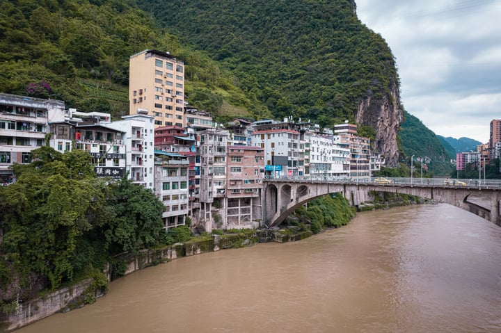 A serene mountain town with white and gray buildings, green trees, and a river flowing under a bridge, surrounded by a mountain and a cloudy sky.