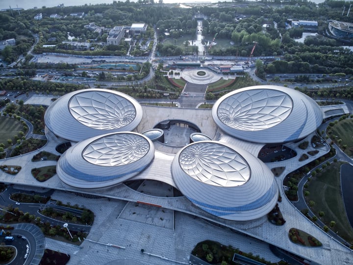 An aerial view of a modern, circular building with four distinct circular structures, surrounded by a large fountain and a park, with a cityscape visible in the background.