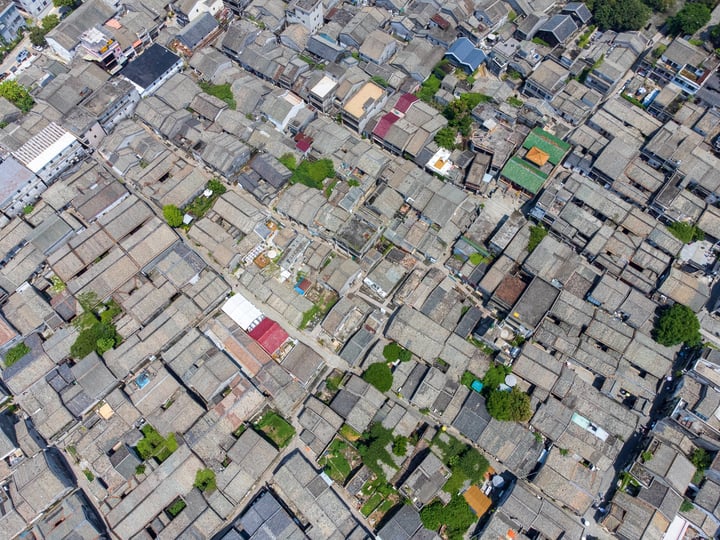 An aerial view of a densely populated city with gray and brown buildings, some with red roofs, surrounded by greenery.