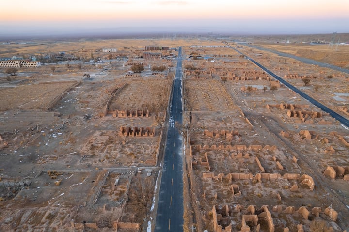 An aerial view of a desert landscape with a long, narrow road, surrounded by large, weathered ruins, and a clear blue sky.