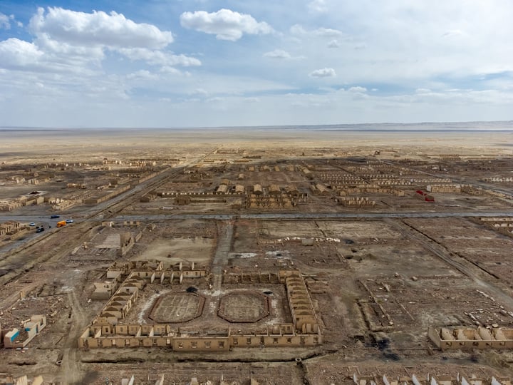 An aerial view of a desert city with a grid-like layout, surrounded by a vast expanse of sand and a clear blue sky with scattered clouds.