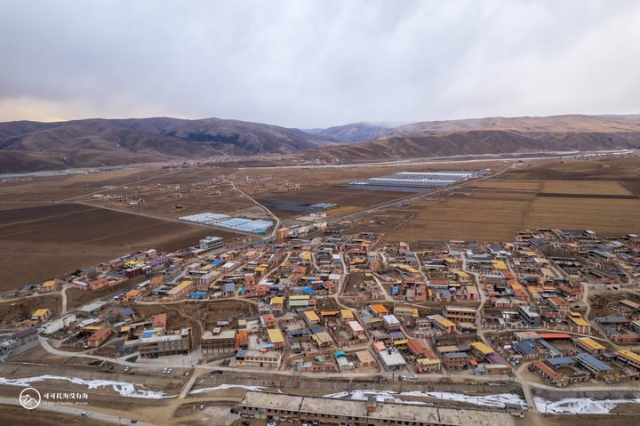 An aerial view of a small town nestled in a valley, surrounded by mountains and surrounded by a vast expanse of barren land. The town is densely packed with houses and buildings, with a few scattered fields and water bodies. The sky is cloudy, casting a soft light over the scene.