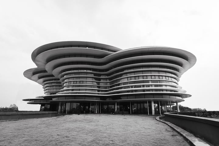 A black and white photograph captures a modern, curved building with a large, curved roof and numerous windows, set against a cloudy sky. The building is surrounded by a grassy area and a concrete wall, with a few people visible in the foreground.