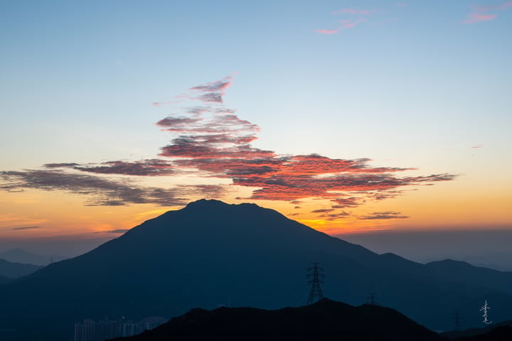 A mountain range with a dark sky and a sun setting behind it, with power lines in the foreground.