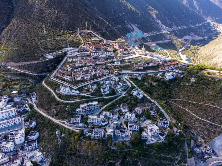 An aerial view of a mountain town with white buildings, red roofs, and a large temple, surrounded by lush greenery and a blue river.