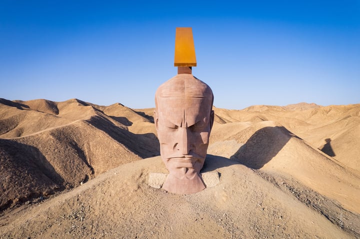 A large, pinkish-brown sand sculpture of a face with a yellow sun on top, standing on a hill in a desert landscape.