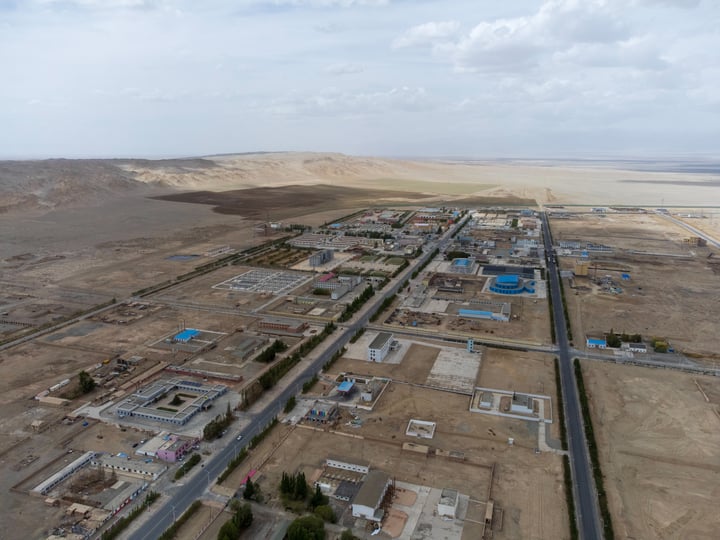 An aerial view of a large construction site with a road, buildings, and a sandy dune in the background.