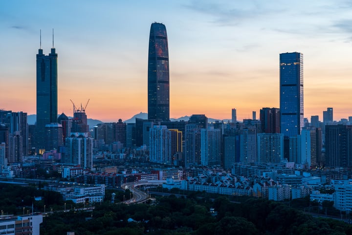 A city skyline at dusk, with a prominent skyscraper, a bridge, and a road, surrounded by buildings of varying heights and colors.