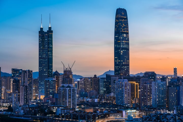 A city skyline at dusk, with a prominent skyscraper, a curved building, and a mountain range in the background.