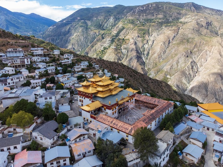 An aerial view of a mountain village with white houses and yellow roofs, surrounded by lush greenery and mountains. The village is nestled on a hillside, with a large temple in the center.