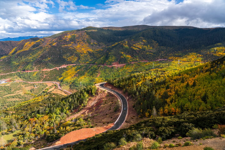A winding black road cuts through a lush valley, surrounded by vibrant autumn foliage. The sky is a clear blue with white clouds, and mountains rise in the distance. The image is taken from a high vantage point, showcasing the vastness of the landscape.