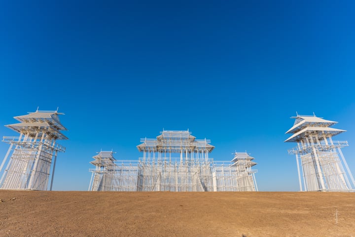 A large, white, glass-enclosed structure with multiple levels stands in a desert-like environment, surrounded by a clear blue sky.