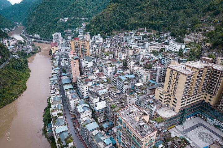 A cityscape on a hillside, with tall buildings of varying heights and colors, surrounded by lush greenery. The river, a brown, choppy body, flows through the city, reflecting the overcast sky.
