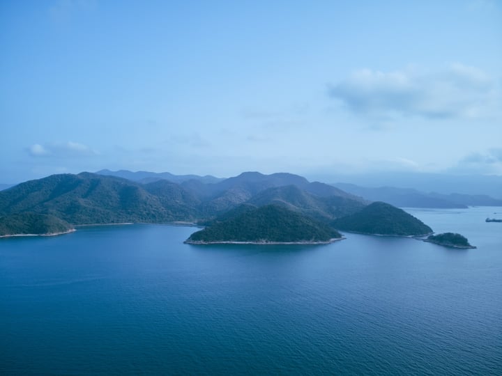 An aerial view of a deep blue ocean with two islands, one on the left and one on the right, surrounded by green hills and mountains. The sky is light blue with a few clouds.