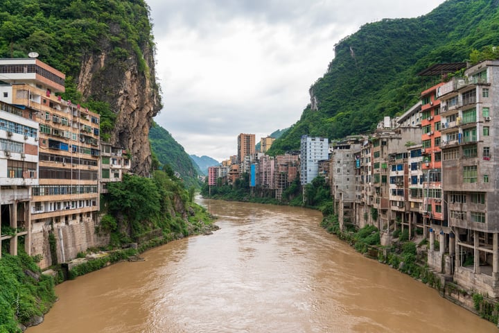 A serene mountain landscape features a winding river, surrounded by colorful buildings on a cliff, with a cloudy sky overhead.