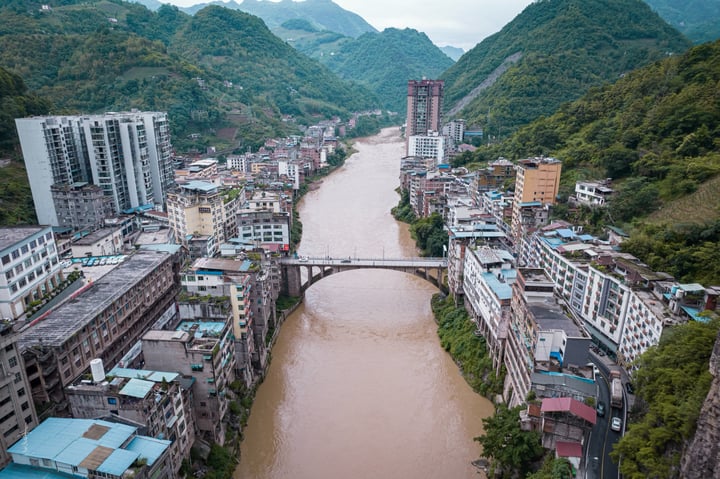 An aerial view of a city with a river, a bridge, and a mix of buildings and trees, surrounded by mountains.