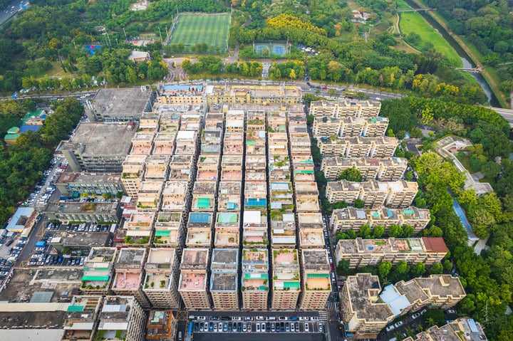 An aerial view of a densely populated urban area with a large grid of buildings, a soccer field, and a park, surrounded by greenery and a road.