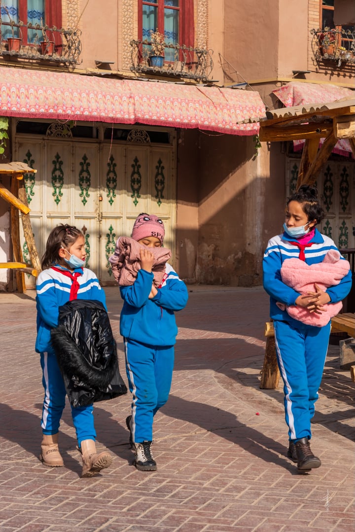 Three children in blue and pink outfits walk down a cobblestone street in a town, with a pink and white striped umbrella in the background.