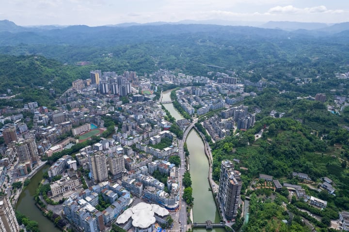 An aerial view of a city nestled between mountains, with tall buildings and a large white dome in the center, surrounded by lush greenery.