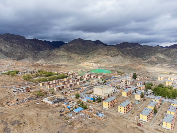 An aerial view of a mountainous area with a construction site in the foreground, surrounded by mountains and a cloudy sky.