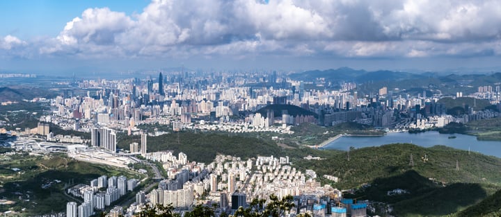 A panoramic cityscape with a mix of tall buildings and a river, surrounded by mountains and greenery, is captured from a high vantage point.
