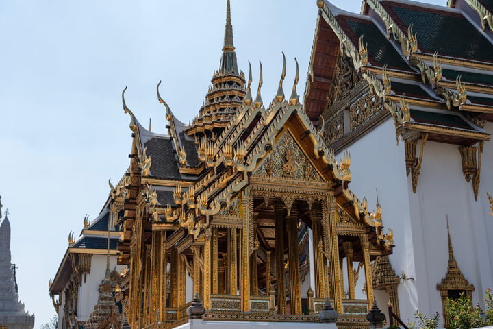 A majestic Thai temple with gold accents and intricate designs stands tall against a clear blue sky, surrounded by lush greenery.
