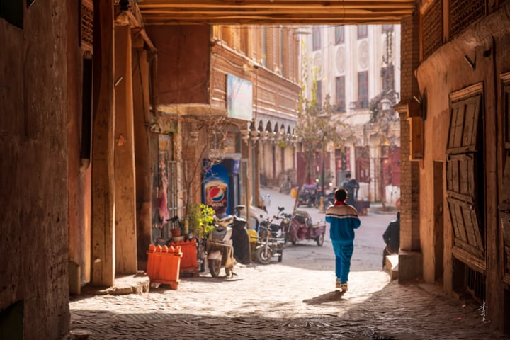 A woman in a blue outfit walks down a narrow alleyway lined with old buildings, surrounded by parked motorcycles and a blue sign. The scene is bathed in sunlight, casting shadows and highlighting the textures of the buildings and the woman's attire.