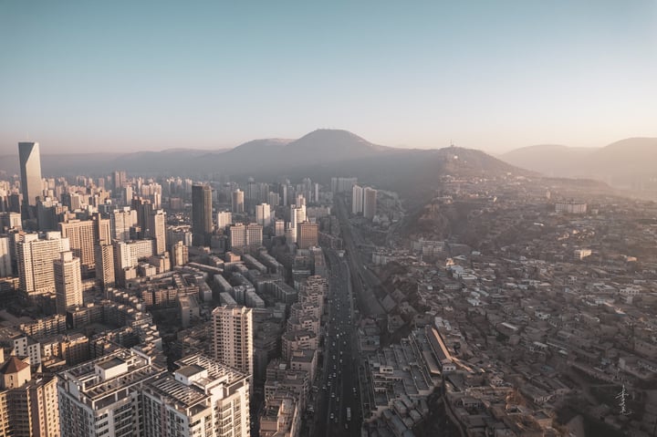 An aerial view of a mountainous city with a hillside, densely populated with buildings of varying heights, surrounded by greenery. The city is situated on a hill, with roads and highways running through it. The sun casts a warm glow on the city, highlighting its architectural details.
