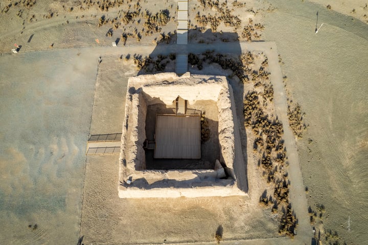 An aerial view of a large, square stone structure surrounded by a grassy area, with a pathway leading to it. The structure has a central entrance and a small roof, and is surrounded by a sandy area. The ground is covered in grass, and the sky is clear.