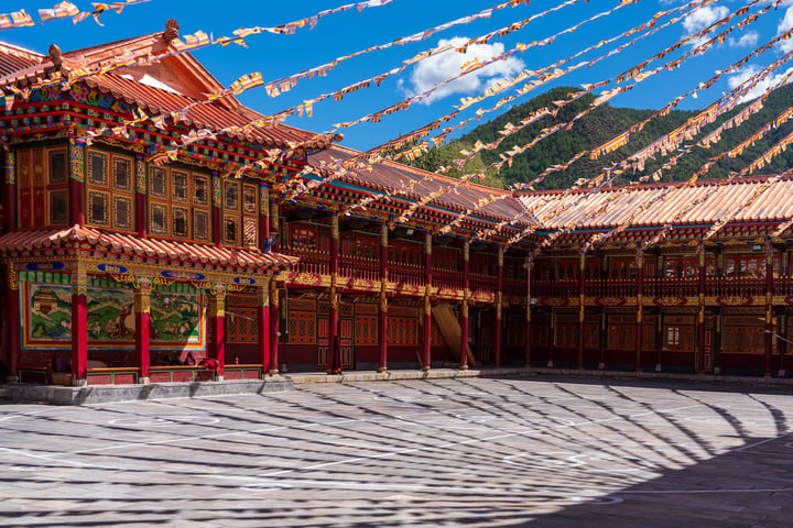 A traditional Chinese temple with red and gold accents, a red roof, and colorful flags is situated in a serene mountainous area. The temple is surrounded by a spacious courtyard with a wooden floor and a large window. The sky is a clear blue with white clouds, and mountains are visible in the distance.